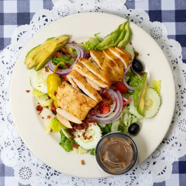 Grilled chicken salad with avocado, bacon, red onion, and olives on a white plate.