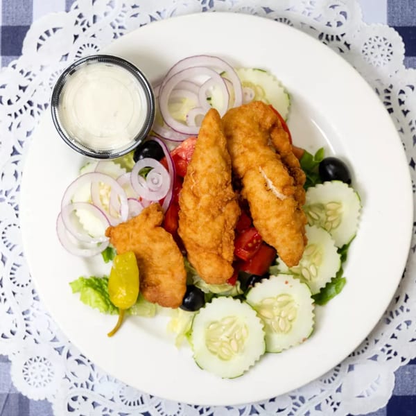 Chicken tender salad with lettuce, tomatoes, cucumbers, onions, olives, and a side of dressing.