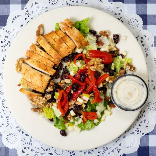Grilled chicken salad with lettuce, red peppers, walnuts, dried cranberries, and dressing on a white plate.
