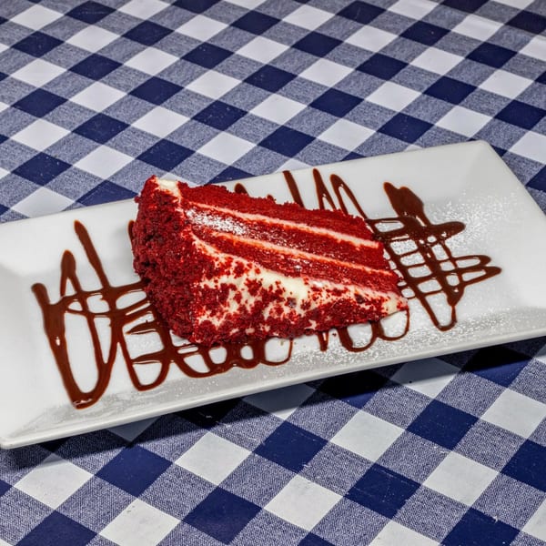 A slice of red velvet cake with cream cheese frosting and chocolate drizzle on a white plate.