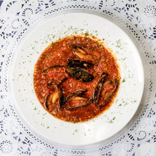 Mussels in a rich red tomato sauce with herbs, served on a white plate with a lace placemat.