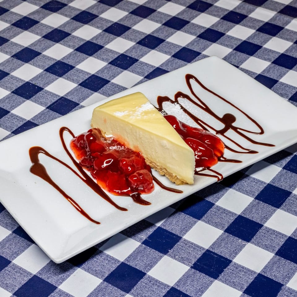 A slice of cheesecake with strawberry topping and chocolate drizzle on a white plate.