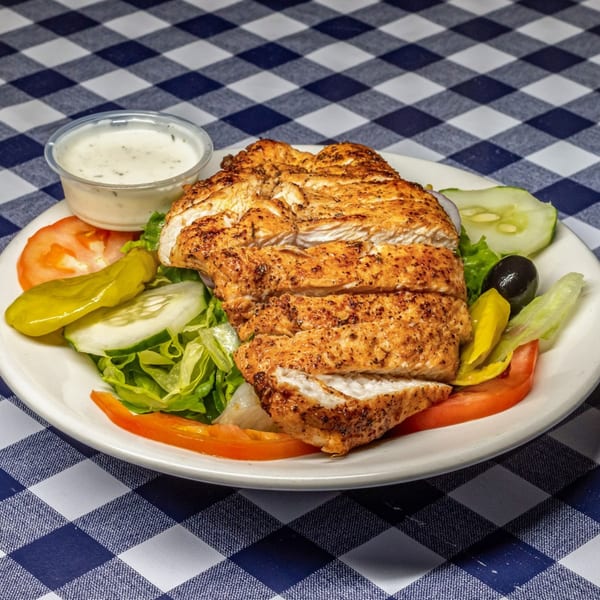 Grilled chicken salad with lettuce, tomatoes, cucumbers, olives, and a side of dressing on a checkered tablecloth.