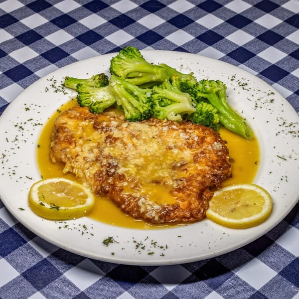 Chicken piccata with lemon slices and broccoli on a white plate, on a blue and white checkered tablecloth.