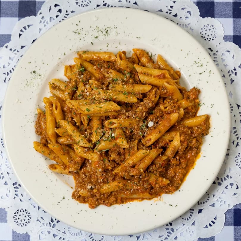 Penne pasta with a rich meat sauce, sprinkled with cheese and herbs, on a white plate.
